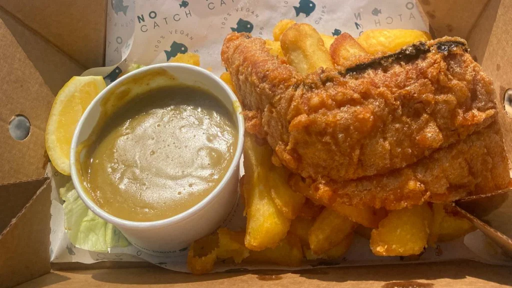 Golden brown battered vegan fish, on top of thick cut chips with a large pot of curry sauce, in a cardboard box