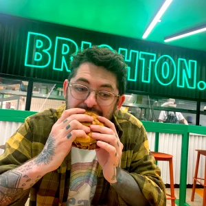 a man eating a burger with a neon sign behind that says 'Brighton'