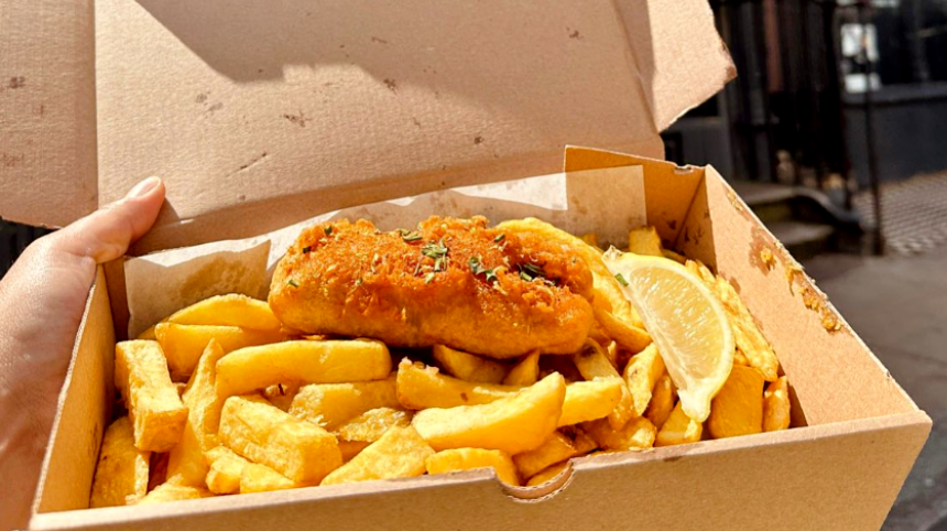 hand holding a cardboard box filled with golden chips and a battered vegan fish with herbs and slice of lemon