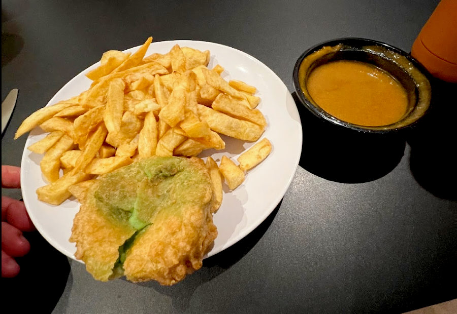 battered pea fritter with chips on a white plate, next to a side of curry sauce on a black table