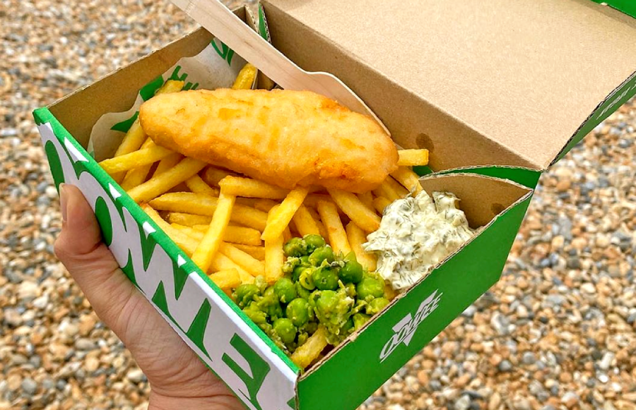 takeaway fish and chips, with tartar sauce and mushy pease, in a cardboard box with a pebbled beach in the background