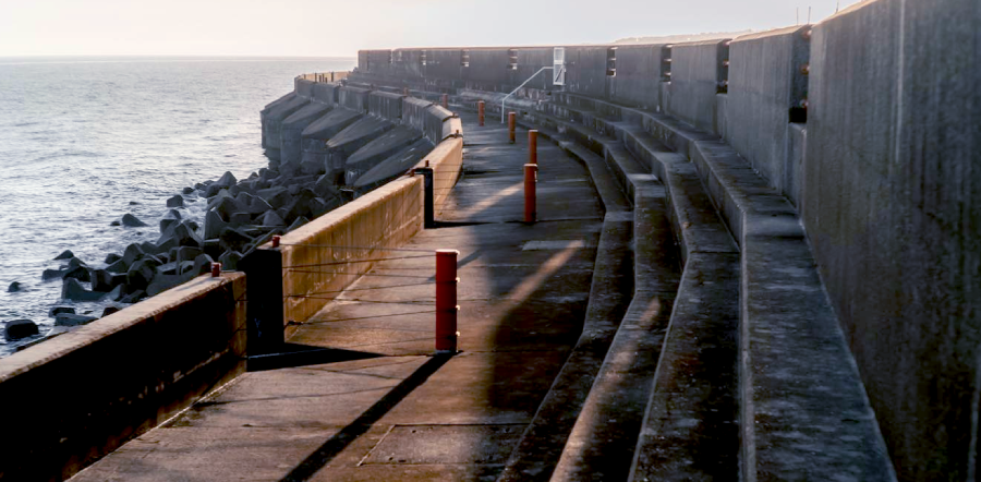 A long concrete marina structure with steps, curving in the distance, by the sea with rocks. A popular fishing spot.