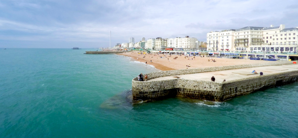 A wide far shot of Brighton Beach. There is a stone and concrete breakwater protruding into the sea. 
