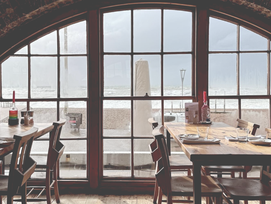 Interior shot of old restaurant with a large brick curved window and several window panes, with tables and chairs. Brighton beach is in the background on a cloudy day.