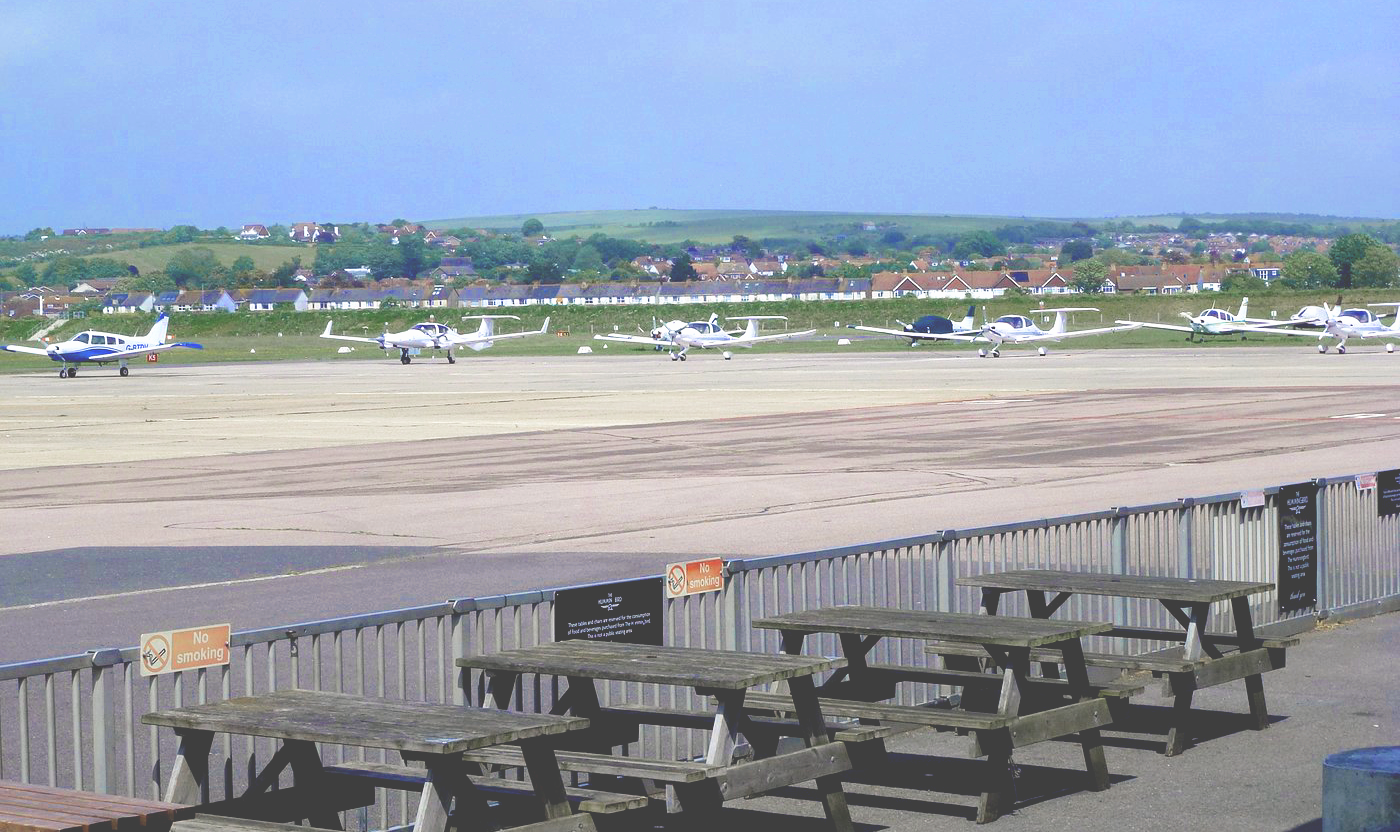A far shot of Brighton airport runway, 6 small propellor planes are parked and in the foreground there are several wooden benches.